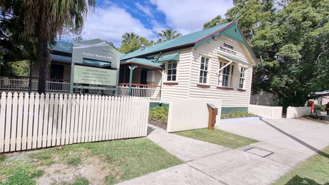 Historic timber building with a green roof and cream picket fence, with signage reading ‘Redcliffe Museum’, located beside a footpath and surrounded by trees.
