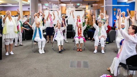 A group of senior people standing in a arc, hands are in the air, a person to the right is showing them what to do.