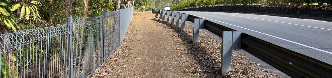 Unsealed roadside walking path bordered by a wire fence and safety guardrail, running alongside a road through a bushland area.