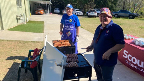 Two people smiling over a BBQ with sausages cooking.