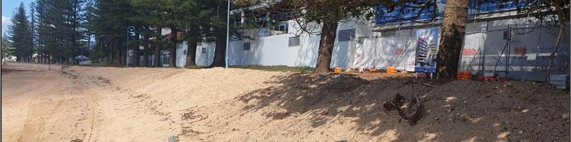 Sandy beach edge with trees and a fenced building area behind, including signage and construction fencing.